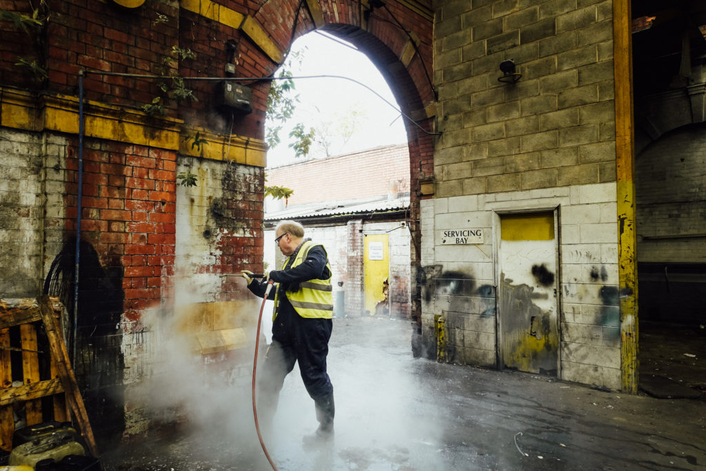 Old stone and brick building with paint and vegetation clinging to the walls being cleaned by man in high vis with steam cleaning 