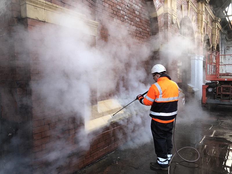 Person in high vis in front of a brick and terracotta wall carrying out steam cleaning with hot steam flowing away.