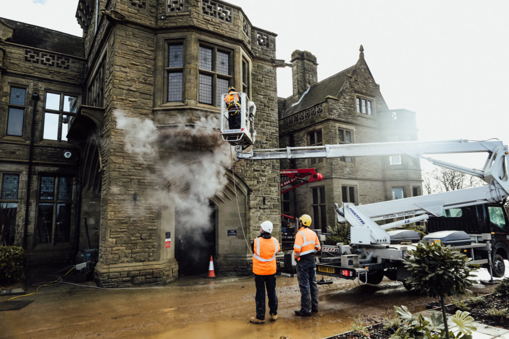 A historic dark stone building being steam cleaned by a person in high vis on a MEWP